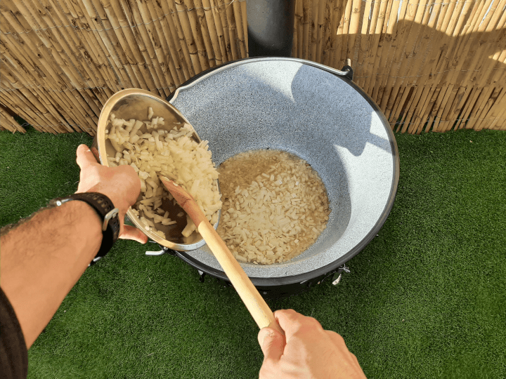la receta del goulash eslovaco a la lena: se ve una mano cogiendo un bol con cebolla picada y otra empujándola a la Kotlina con una cuchara de madera.