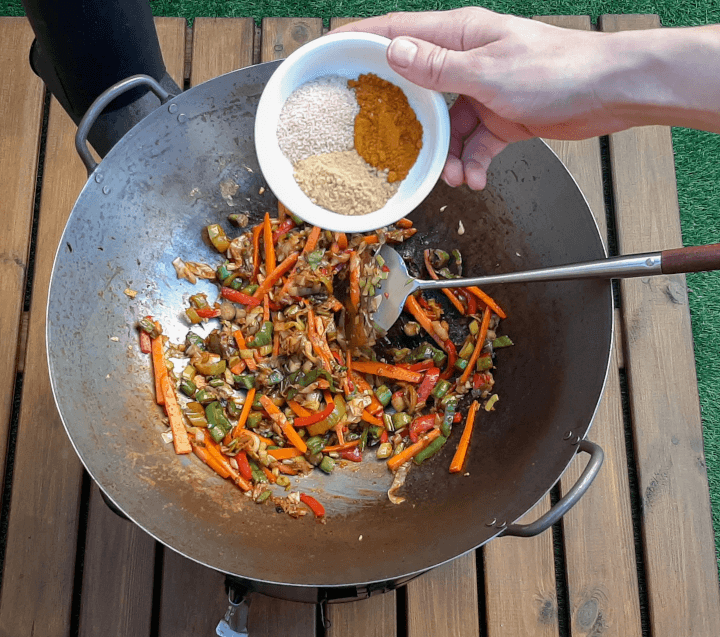 el wok con las verduras ya doradas y una mano echando especias