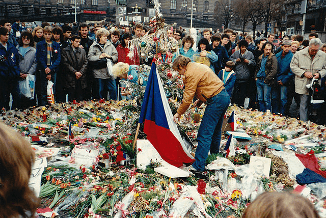 vaclav havel de pie en el centro de un circulo hecho de flores y ofrendas en memoria a las victimas del comunismo rodeado por otros ciudadanos unidos en la protesta