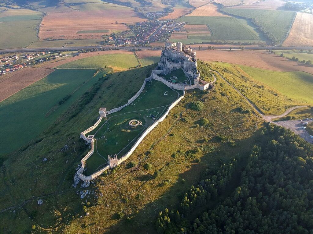 vista aerea del impresionanete castillo de spis en que se ve el castillo en si a lo alto de una colina y la zona amurallada cayendo a la izquierda del lector rodeado de prados verdes