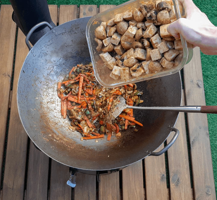 el wok con las verduras y especias y una mano echando el tofu y las setas marinadas