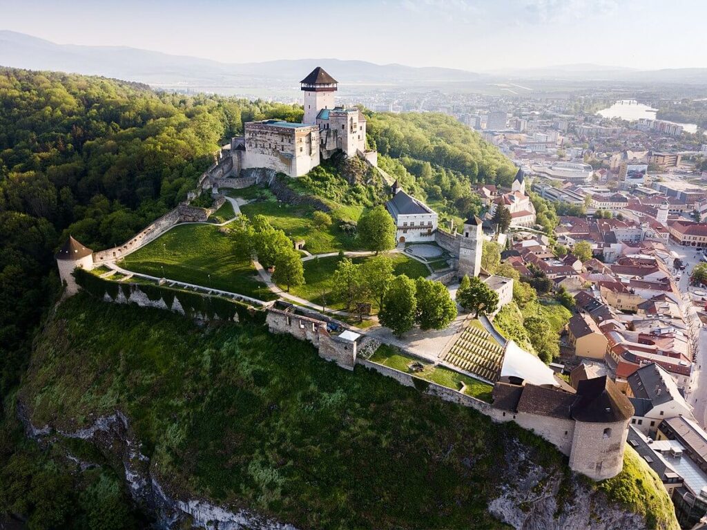 vista aerea del impresionanete castillo de trencin en que se ve el castillo en si a lo alto de una colina en primer plano y un bosque y la ciudad de trencin alrededor
