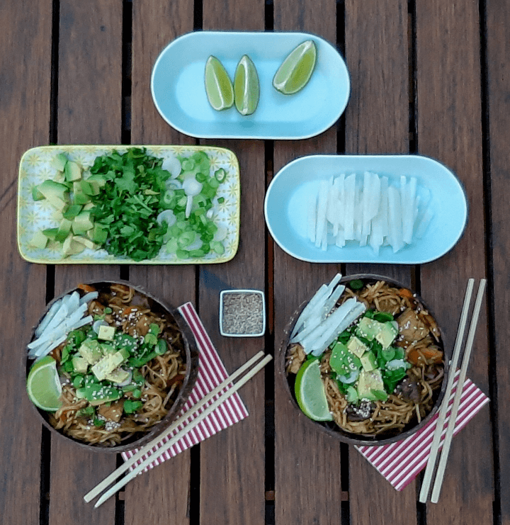 Una mesa de madera en la que hay dos platos de coco con los fideos con verduras y tofu preparados en el wok y tres platillos mas con ingredientes para acompañar como lima, aguacate y cebollino