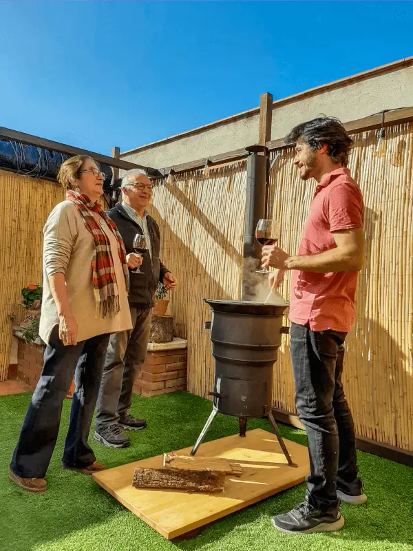 un chico y sus padres de pie al lado de una cocina barbacoa y horno de lena portatil para exterior o kotlina mientras toman una copa de vino