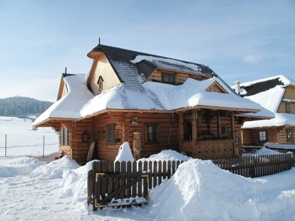 Una Chata o cabana de madera en un paisaje nevado. En la fachada hay una estatua también de madera y un porche pequeño. La casa está rodeada de una valla de madera.