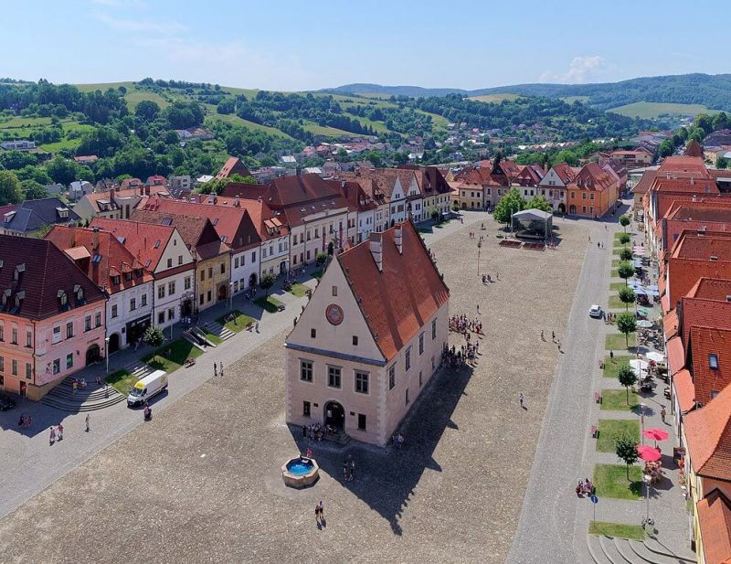 vista aerea de la plaza principal de bardejov en que se ve el ayuntamiento en el centro, y los demas edificios haciendo un rectangulo alrededor. en el segundo plano se ven colinas verdes con arboles y un cielo azul y sin nubes