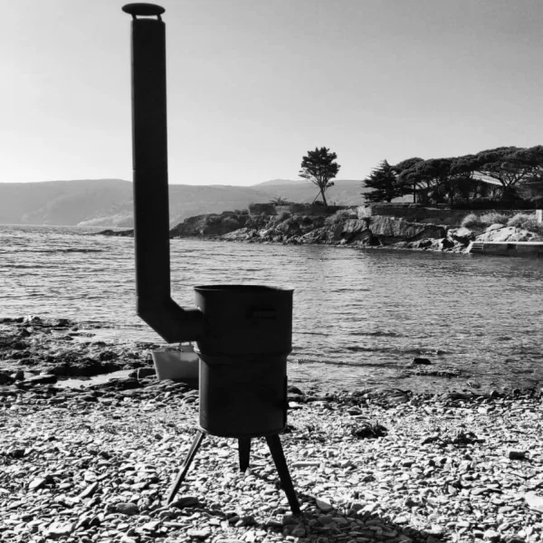 foto en blanco y negro de una kotlina o cocina de leña portátil tipo barbacoa para exterior tradicional de lena en una playa de piedras en cadaques