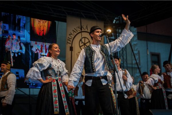 tradiciones y folklore de eslovaquia: una pareja de baile en sus vestidos tradicionales de color blanco y azul saludan al publico desde el escenario mientras en segundo plano el resto de parejas cantan